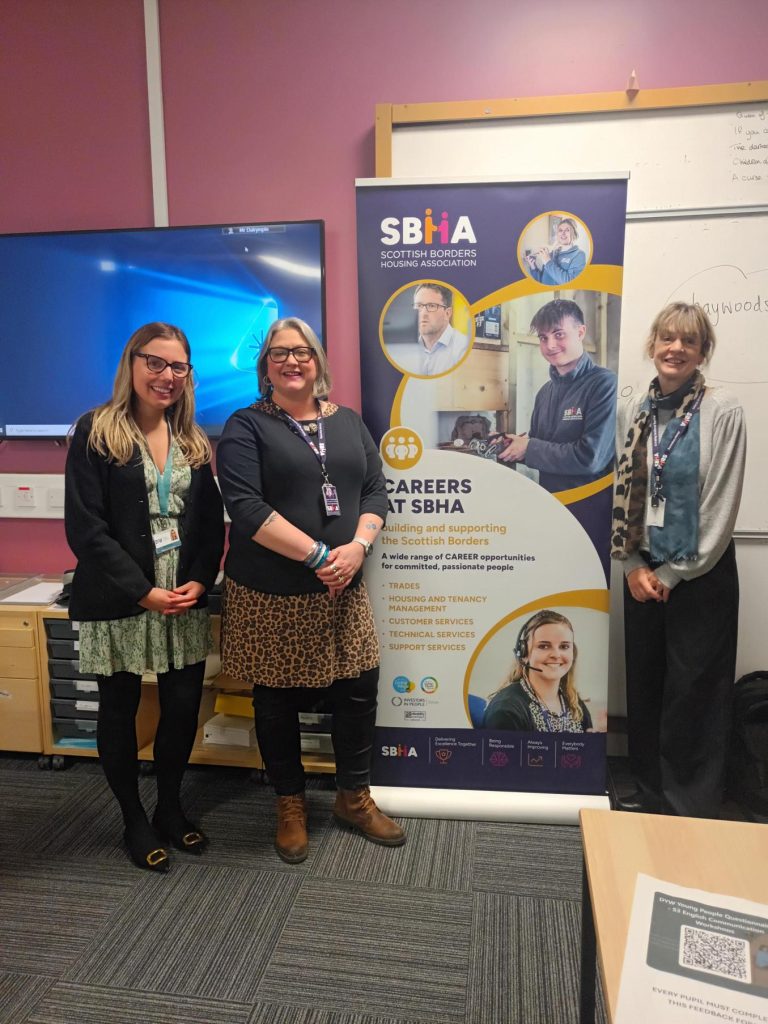 Three women standing in front of a roll-up banner for the Scottish Borders Housing Association (SBHA).