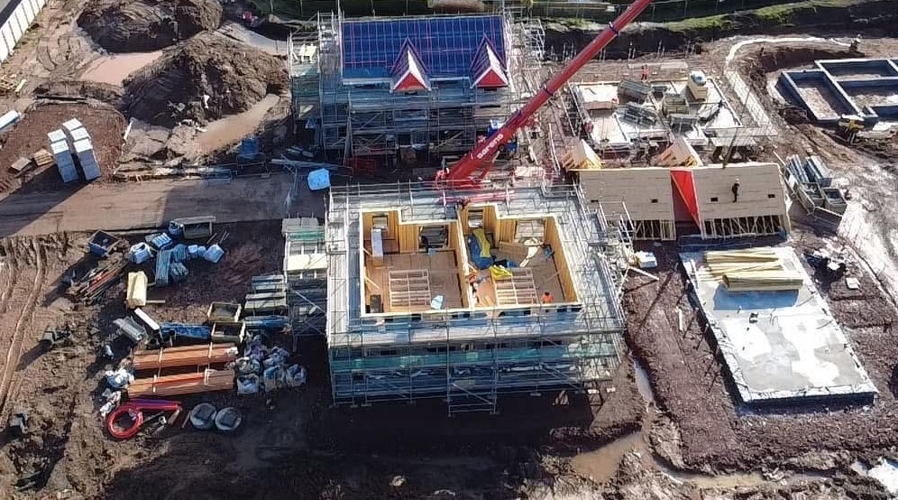 Aerial view of a construction site featuring a partially built house with wooden framework and scaffolding. A red crane is positioned over the structure, lifting building materials. The roof frame with red and white triangular gables is visible, and workers can be seen on the site. Surrounding the main structure are piles of building materials, concrete foundations, and muddy terrain. Several smaller structures and construction equipment are scattered throughout the area.
