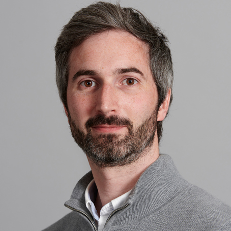 Man with short hair and a beard, wearing a light gray zip-up sweater over a white collared shirt, posing in front of a plain gray background.