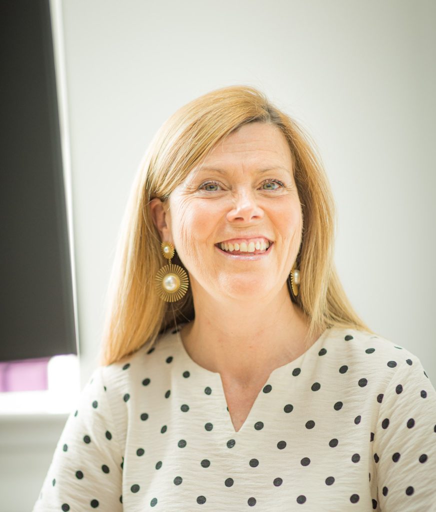 A smiling woman with blonde hair sits indoors, wearing a cream blouse with black polka dots and large statement earrings.