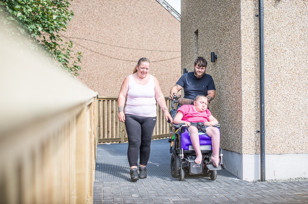 A woman and man accompany a child using a powered wheelchair along an accessible ramp beside a building. The woman walks alongside while the man guides the wheelchair, with the child seated in the chair.