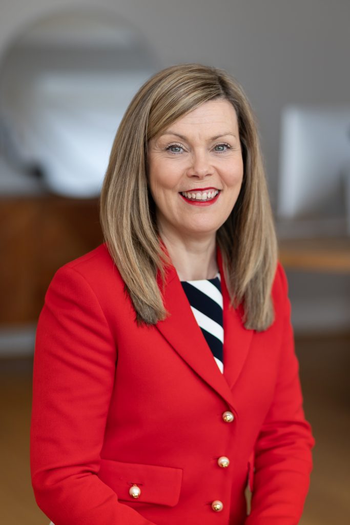 A smiling woman with shoulder-length blonde hair wearing a bright red blazer and striped top, posing indoors against a softly blurred background.