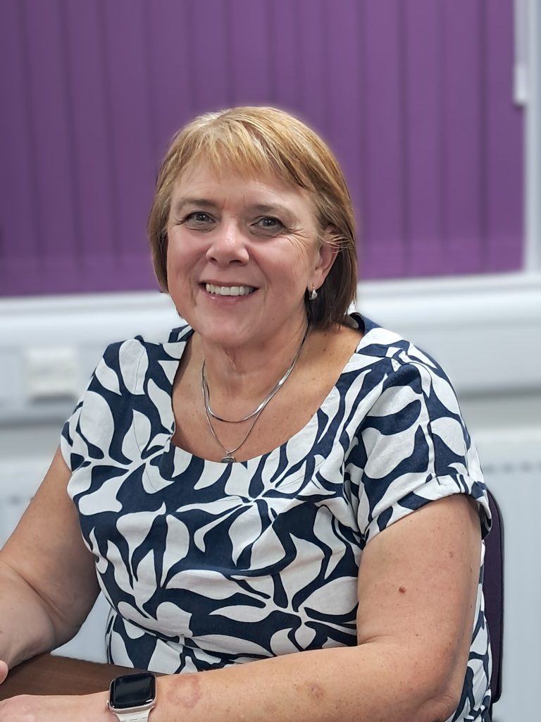 A smiling woman with short blonde hair sits at a table, wearing a navy and white patterned top, necklace and smartwatch. She is facing the camera against a purple and white background.