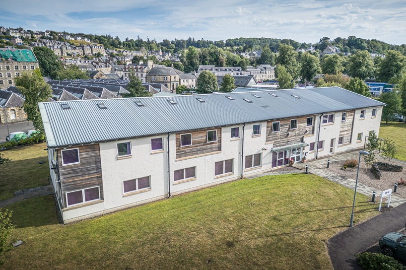 Aerial photo of a modern two-story building with partial timber cladding. A hilly townscape appears in the background.