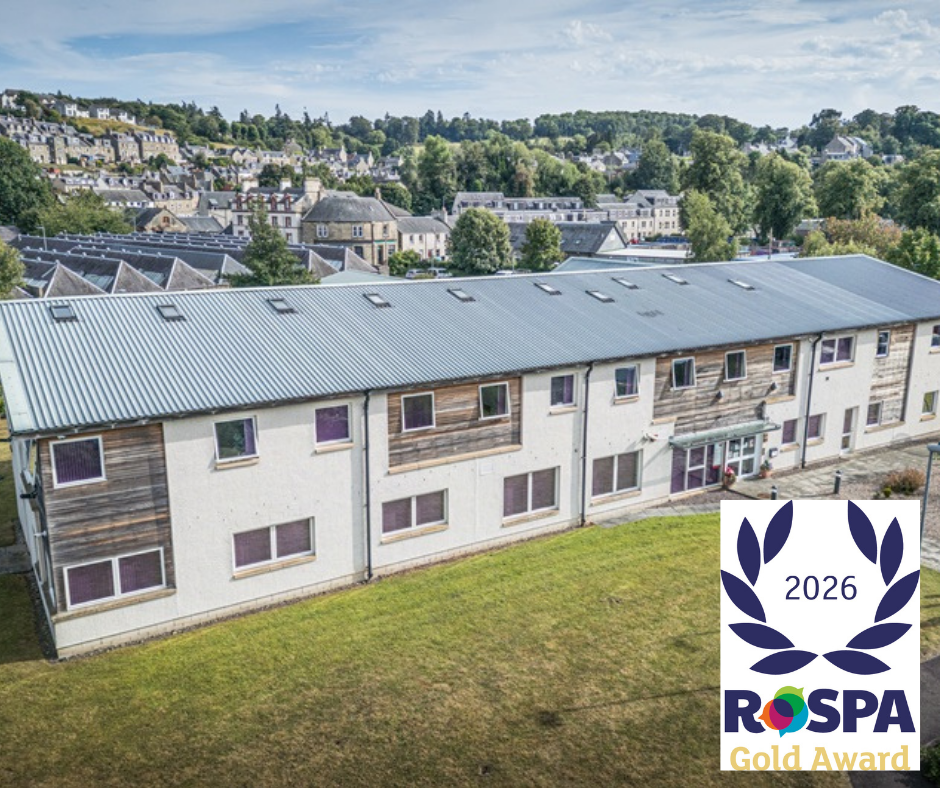 Aerial view of a two-storey housing building with a large grassed area in front, set against a hillside town. A graphic in the bottom right displays the RoSPA 2026 Gold Award.